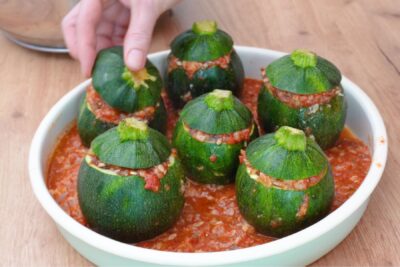 Dovlecei rotunzi umpluți cu orez și ciuperci, așezați într-o tavă cu sos de roșii, gata de copt la cuptor