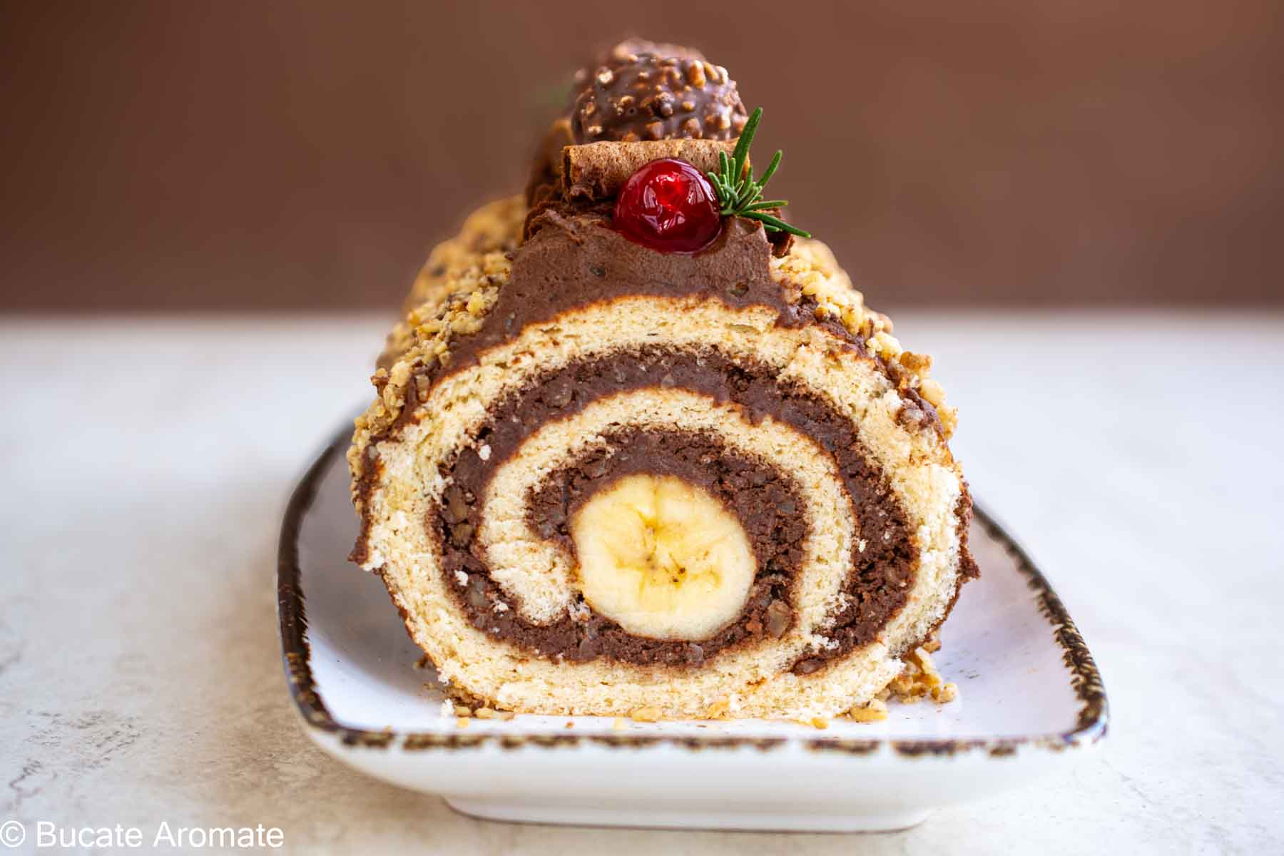 Ruladă cu cremă de ciocolată și banană în mijloc, decorată cu crocant de nuci – felie văzută în secțiune. Titlu SEO: