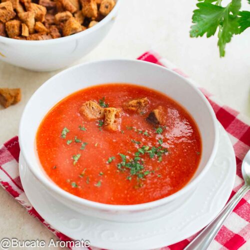 Supă cremă de ardei copți servită în bol alb, cu crutoane crocante și pătrunjel verde deasupra