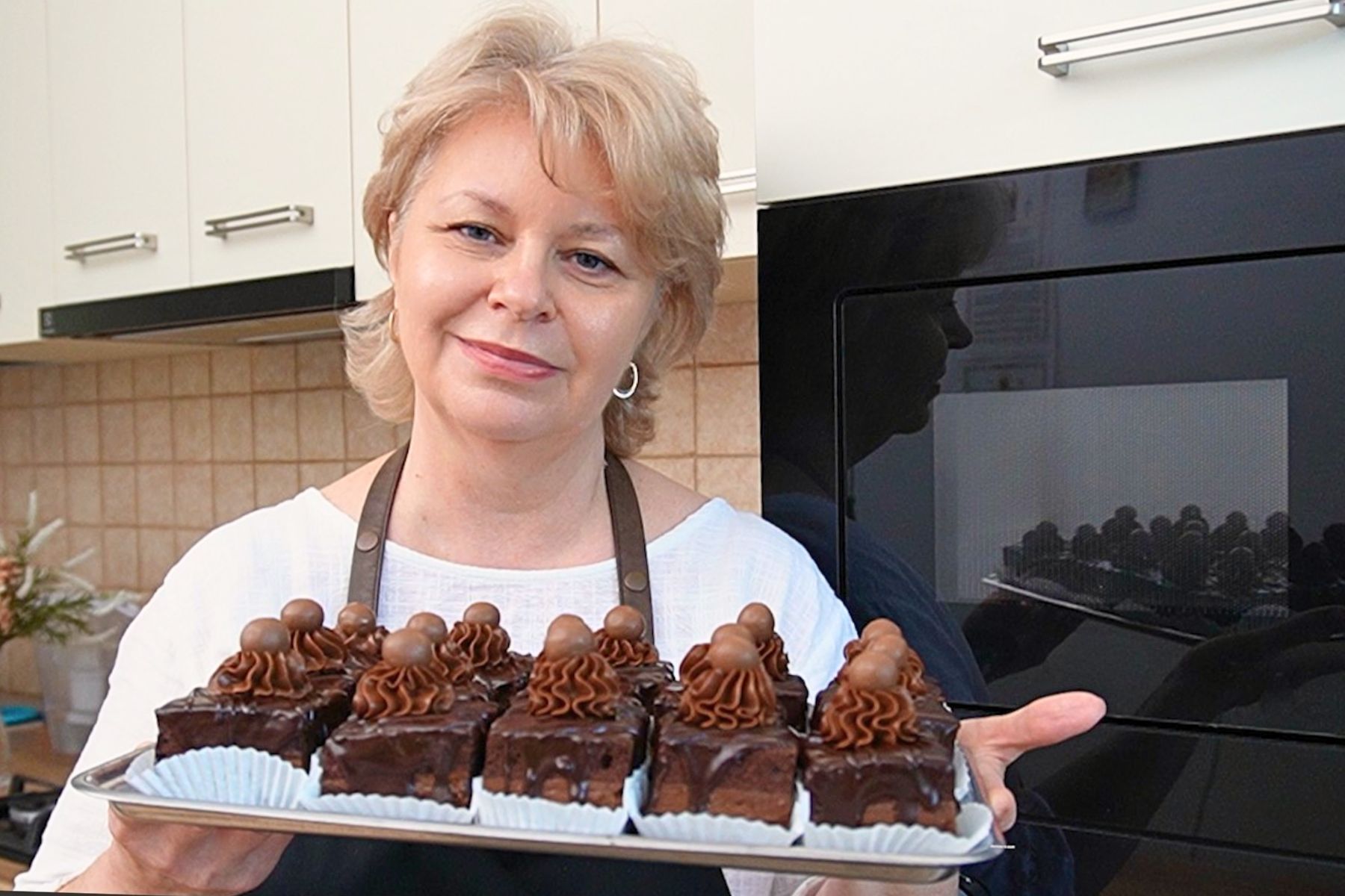 Iuliana Sbirnea ținând în mână o tavă cu amandine de casă cu ciocolată, decorate cu ganache și glazură lucioasă, în bucătărie