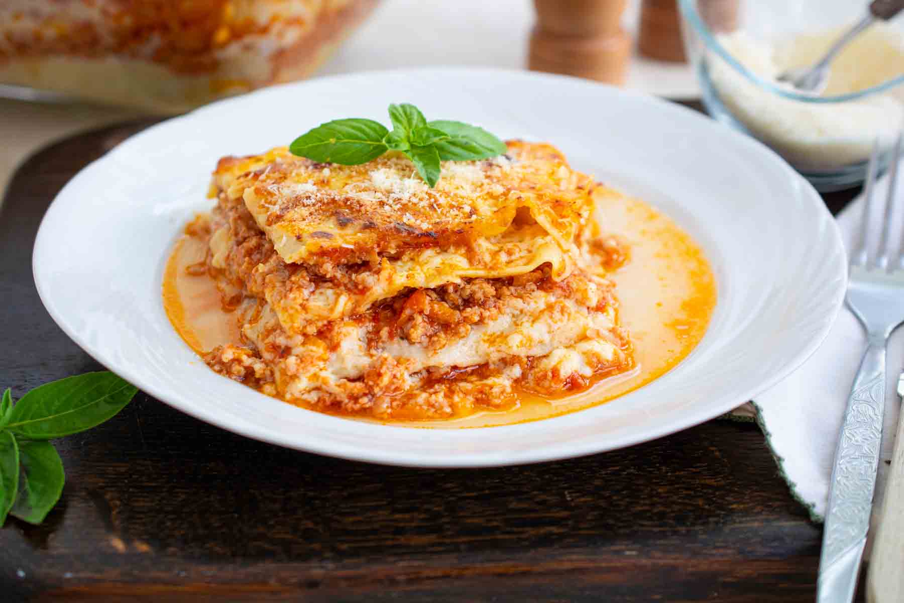 o portie de lasagna bolognese pe farfurie alba decorata cu busuioc