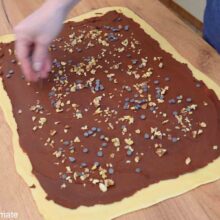 Aluat de Babka întins pe blat, acoperit cu umplutură de ciocolată și presărat cu nuci și picături de ciocolată