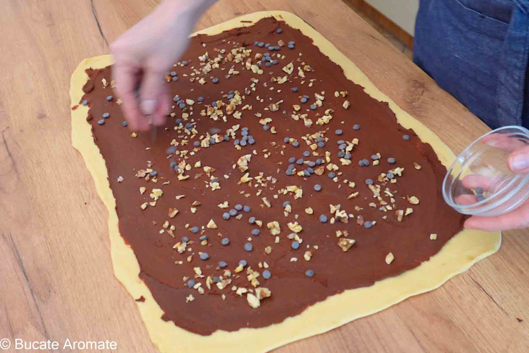 Aluat de Babka întins pe blat, acoperit cu umplutură de ciocolată și presărat cu nuci și picături de ciocolată