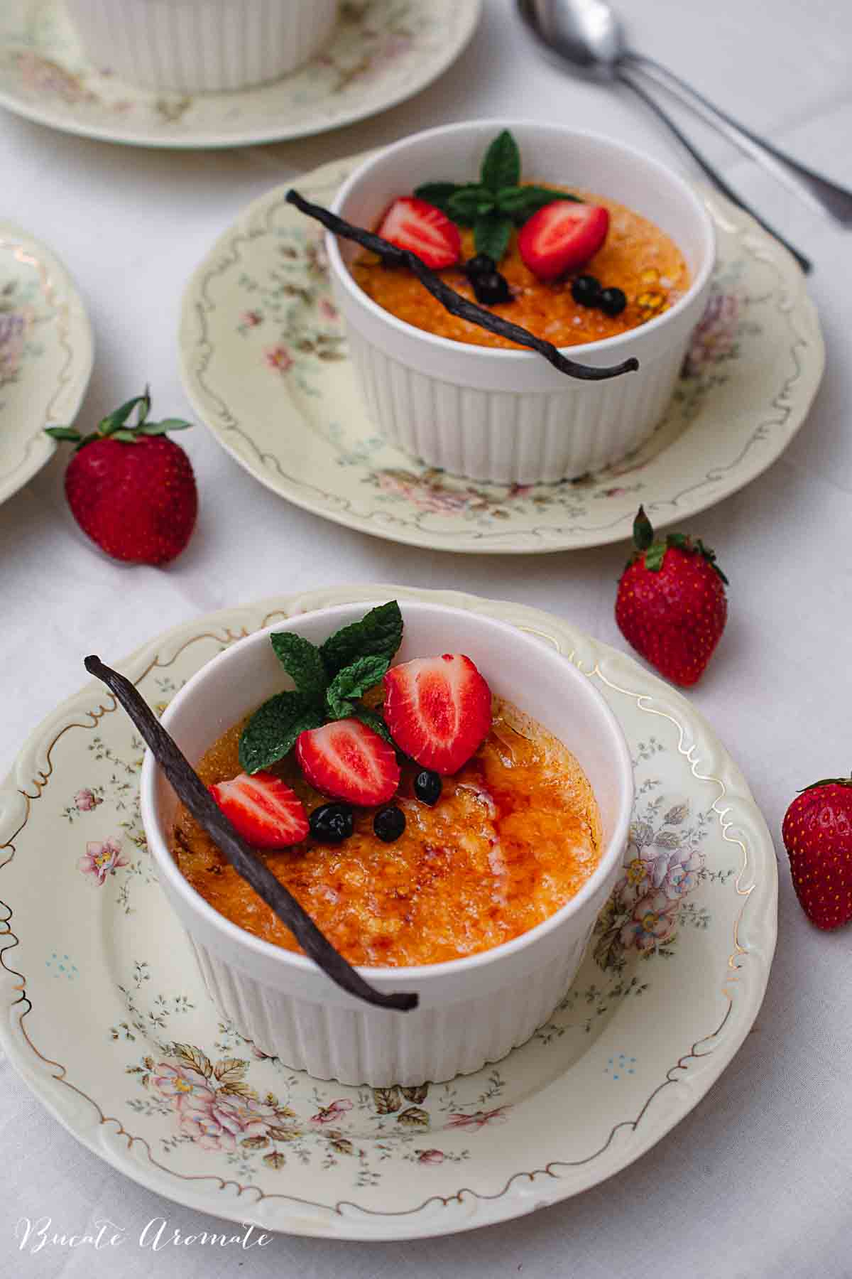Două porții de crème brûlée cu crustă caramelizată, decorate cu căpșuni, afine, mentă și păstaie de vanilie.