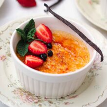 Crème brûlée servită în ramekin, decorată cu căpșuni, afine, mentă proaspătă și o păstaie de vanilie.