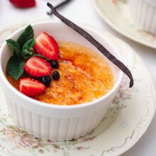 Crème brûlée cu crustă crocantă de zahăr, căpșuni proaspete, afine, mentă și păstaie de vanilie, servită în ramekin pe farfurie vintage.