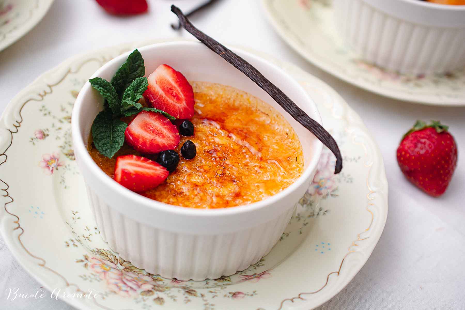 Crème brûlée cu crustă crocantă de zahăr, căpșuni proaspete, afine, mentă și păstaie de vanilie, servită în ramekin pe farfurie vintage.