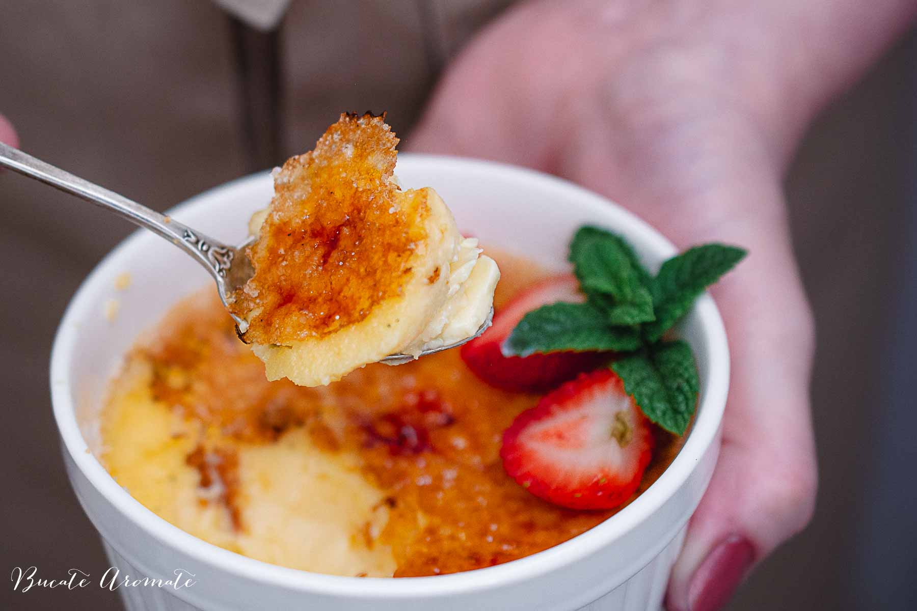 Linguriță cu cremă de vanilie și crustă caramelizată, luată dintr-o porție de crème brûlée decorată cu căpșuni și mentă.