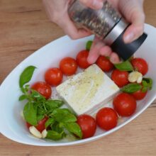Condimentarea amestecului de roșii cherry, brânză feta, usturoi și busuioc cu piper proaspăt măcinat, înainte de coacere.