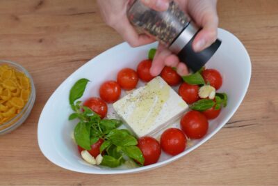 Condimentarea amestecului de roșii cherry, brânză feta, usturoi și busuioc cu piper proaspăt măcinat, înainte de coacere.