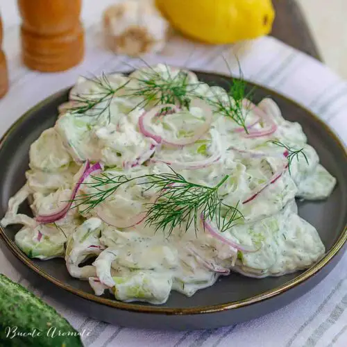 Salată de castraveți cu sos cremos de iaurt și mărar, decorată cu rondele de ceapă roșie și mărar verde.