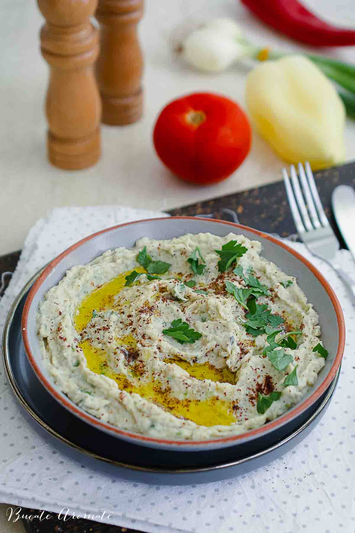Pastă tartinabilă de vinete cu tahini, ulei extravirgin și sumac, pe fundal cu roșii și ceapă verde.