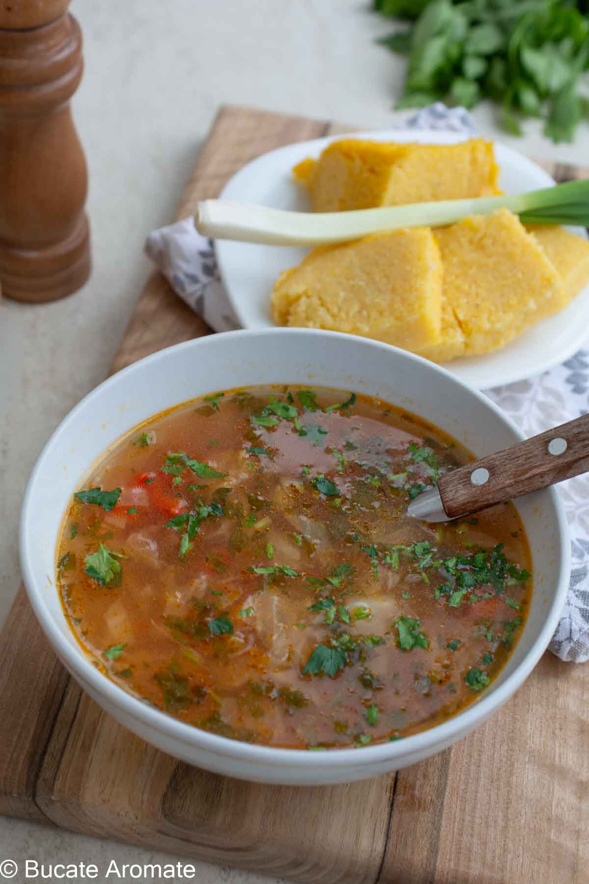 Bol cu ciorbă de varză albă de post, cu borș, orez și legume, servită cu mămăligă caldă și verdeață tocată.