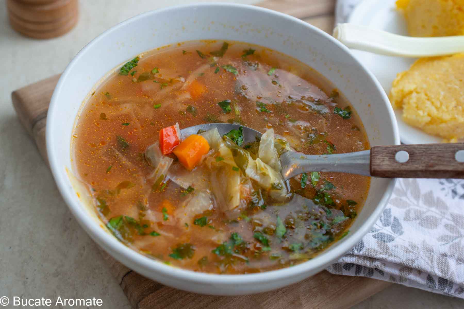 Lingură cu ciorbă de varză de post, cu bucăți de varză, morcov, ardei și verdeață, într-un bol cu zeamă acrișoară.