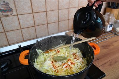 Turnare apă fierbinte peste varza tocată cu boia și dafin, în prepararea ciorbei de varză dulce de post cu borș.