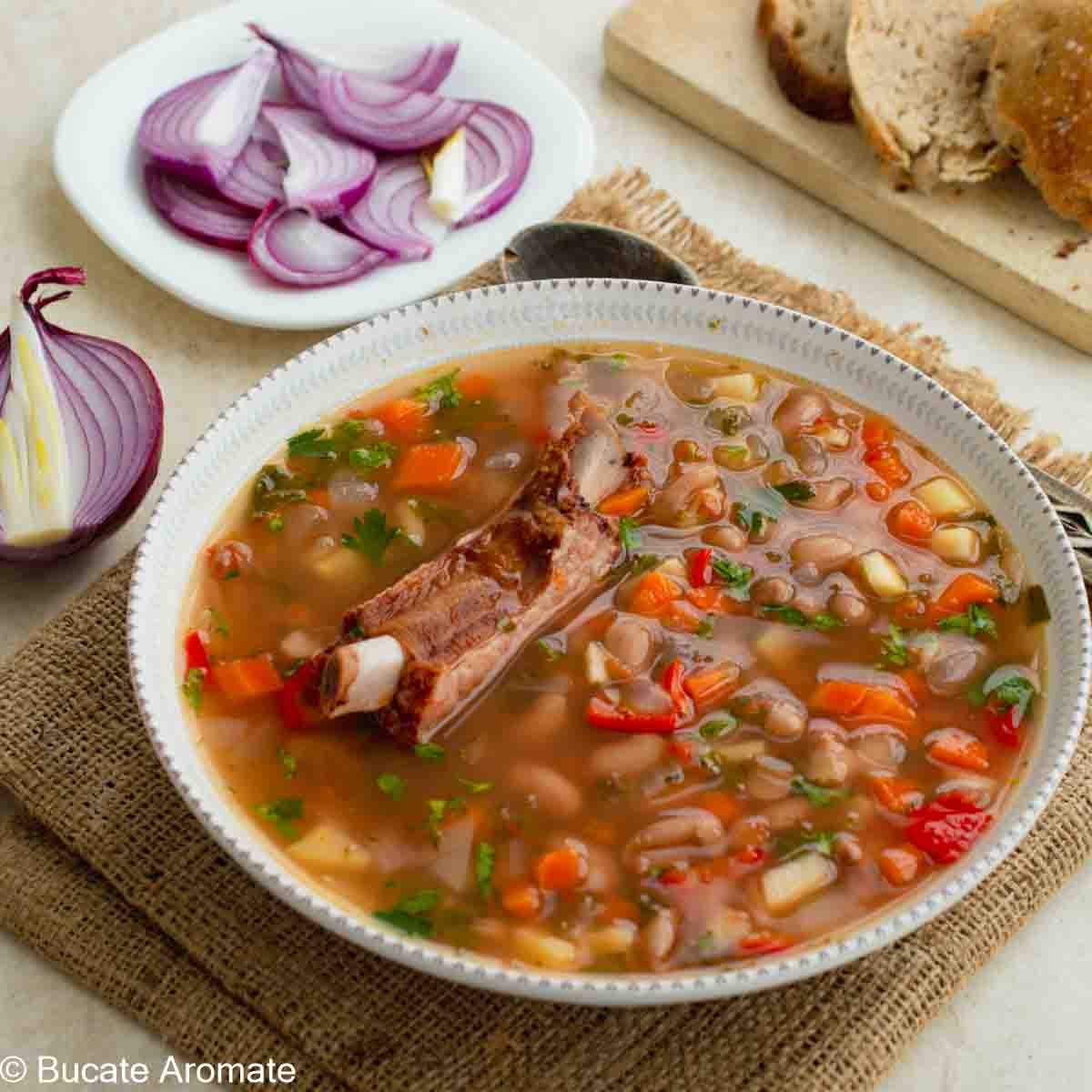 Ciorbă de fasole cu costiță afumată și multe legume servită cu pâine proaspăta și ceapă roșie