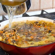 Supă fierbinte de legume turnată peste orez, ciuperci și morcov în tigaie, înainte de coacere, pentru orez cu ciuperci de post la cuptor