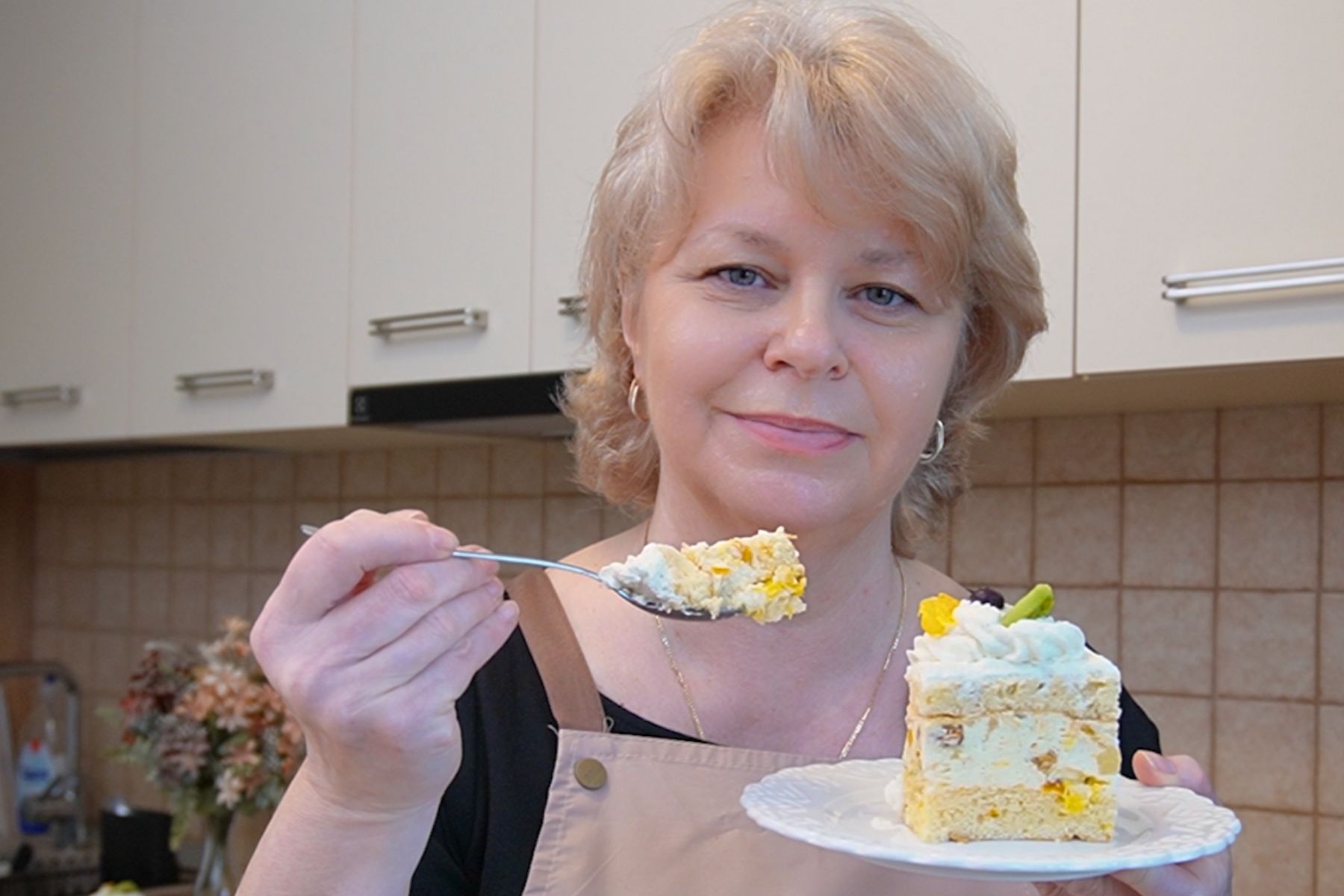 Felie de prăjitură Diplomat la tavă, ținută pe farfurie, în timp ce gust din ea cu lingurița; se văd blatul pufos, crema stabilă și fructele din interior