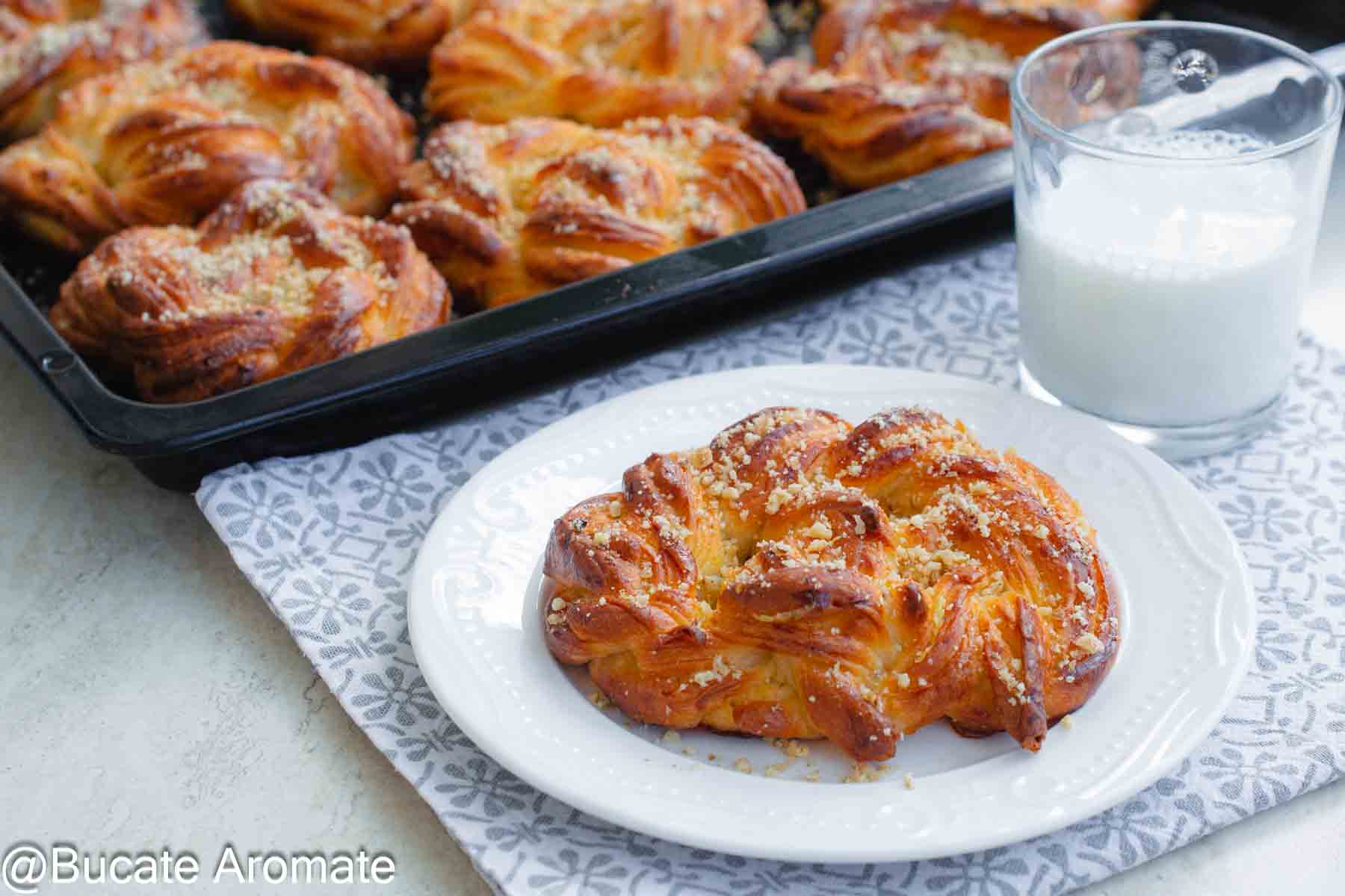 Covrig polonez însiropat servit pe farfurie albă, alături de un pahar cu lapte, cu tava în fundal.