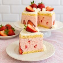 Felie de tort cu spumă de căpșuni, blat de pandișpan și bucăți de căpșuni în cremă, servită pe farfurie, cu tortul întreg în fundal