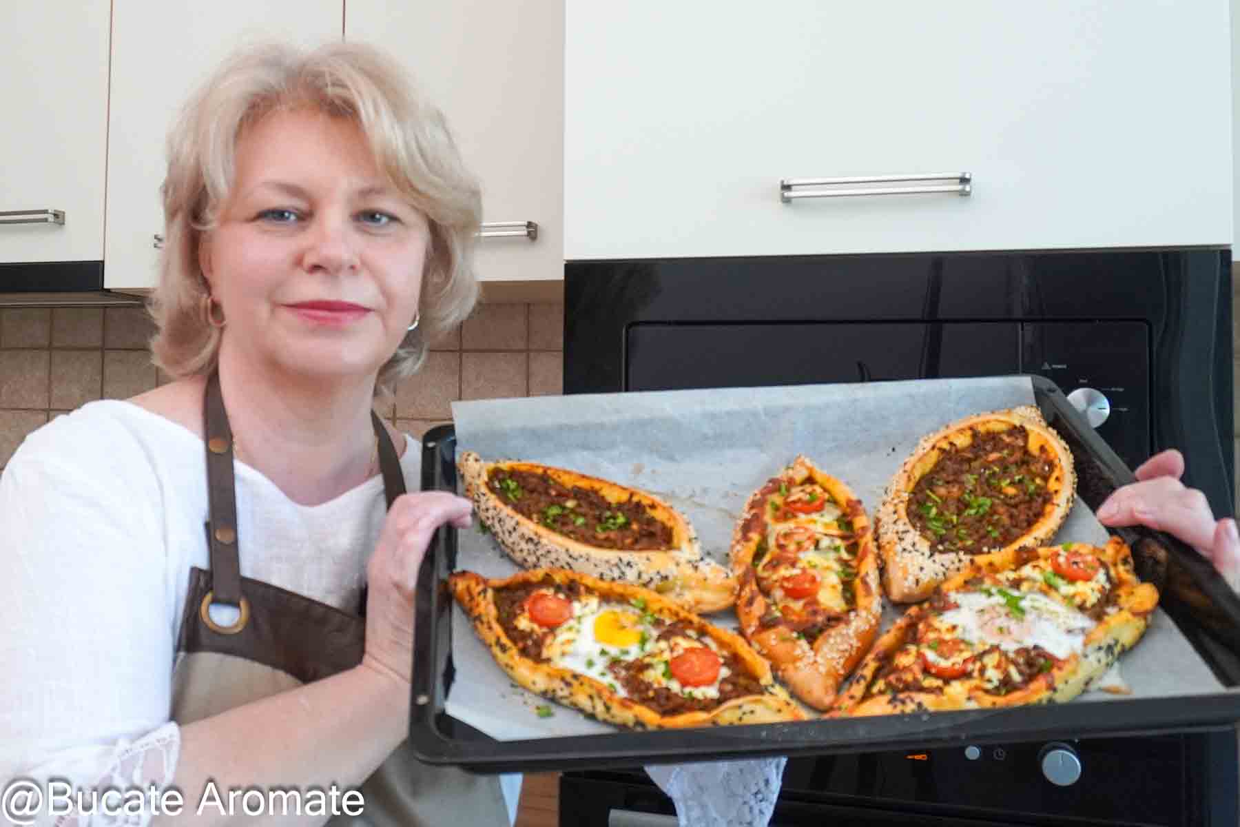 Iuliana de la Bucate Aromate ține o tavă cu pide turcești cu carne tocată de vită, ou, telemea și roșii cherry.