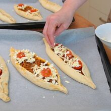 Pide turcești cu carne tocată de vită decorate cu telemea, cașcaval ras și roșii cherry înainte de coacere.