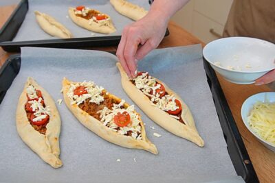 Pide turcești cu carne tocată de vită decorate cu telemea, cașcaval ras și roșii cherry înainte de coacere.