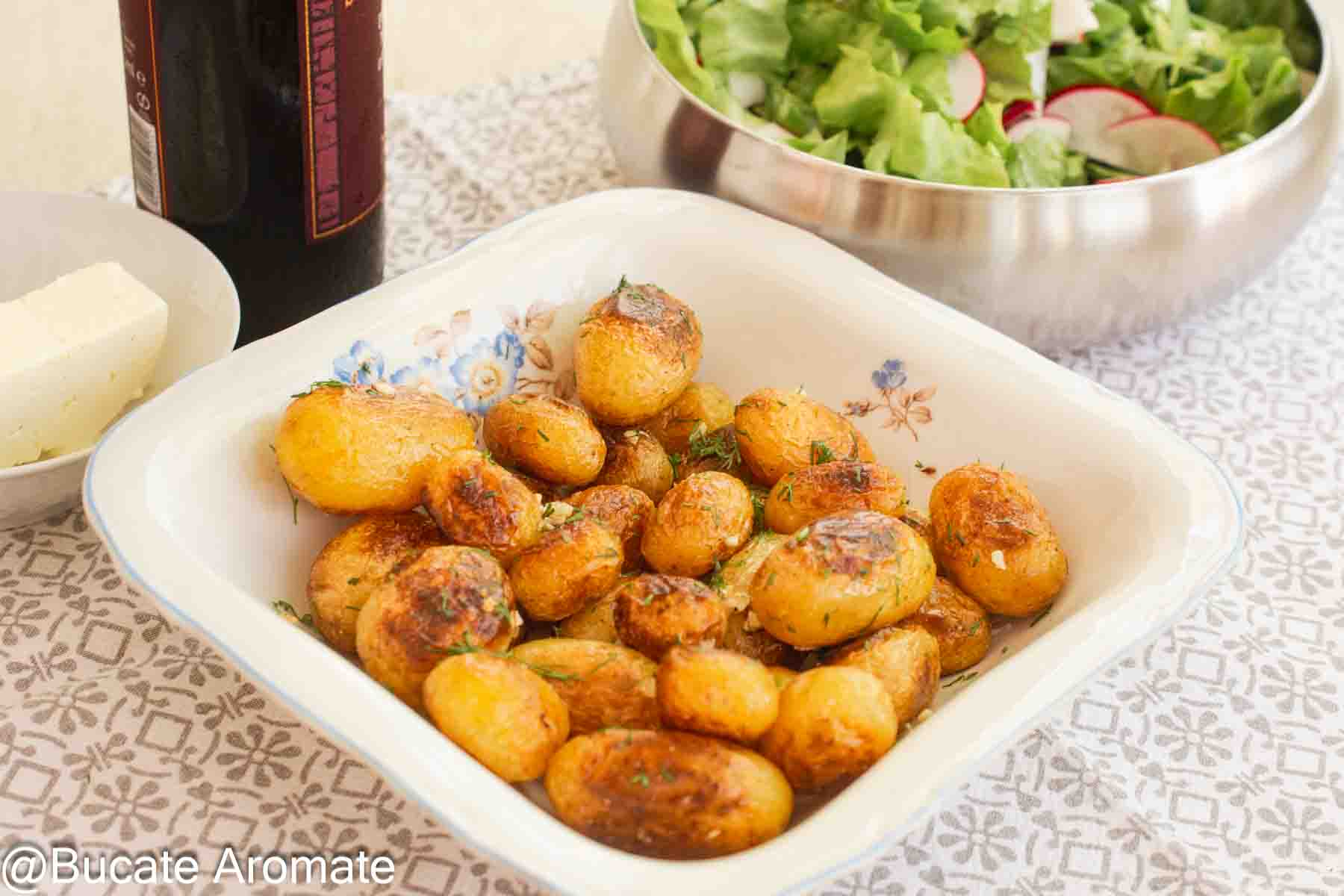 Cartofi noi la tigaie, rumeniți cu unt, ulei, usturoi și mărar, serviți într-un vas alb alături de salată verde.