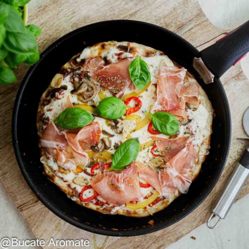 Pizza la tigaie fără drojdie, cu blat subțire, cașcaval topit, prosciutto crudo, ardei, ciuperci și frunze de busuioc.