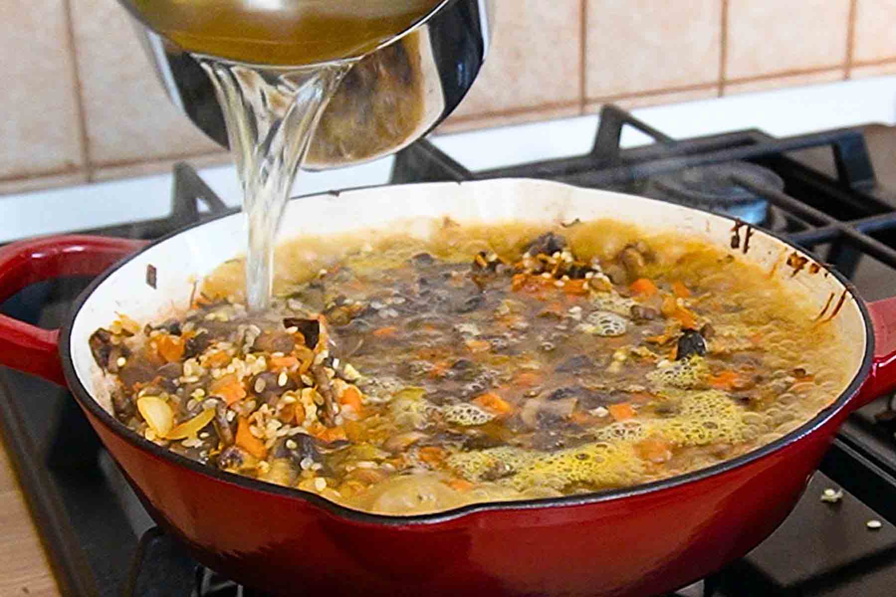 Supă fierbinte de legume turnată peste orez, ciuperci și morcov în tigaie, înainte de coacere, pentru orez cu ciuperci de post la cuptor