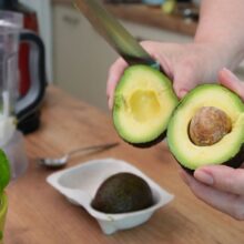 Avocado copt tăiat în jumătate pentru supă rece de avocado cu iaurt, lângă busuioc proaspăt și blender