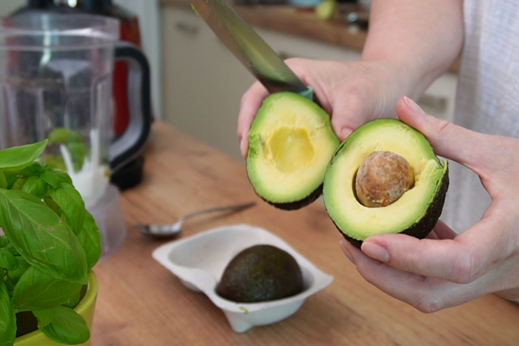 Avocado copt tăiat în jumătate pentru supă rece de avocado cu iaurt, lângă busuioc proaspăt și blender