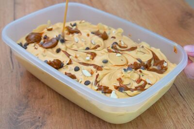 Înghețată cu dulce de leche decorată în caserolă cu dungi de caramel, arahide tocate și picături de ciocolată.