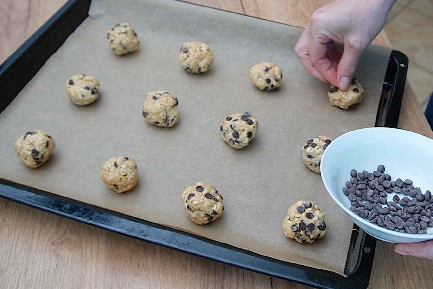 Porții de aluat pentru biscuiți cu ovăz așezate pe hârtie de copt și decorate cu picături de ciocolată
