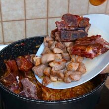 Bucăți rumenite de spată de porc și oase garf afumate adăugate peste varza dulce din cratiță.