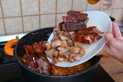 Bucăți rumenite de spată de porc și oase garf afumate adăugate peste varza dulce din cratiță.