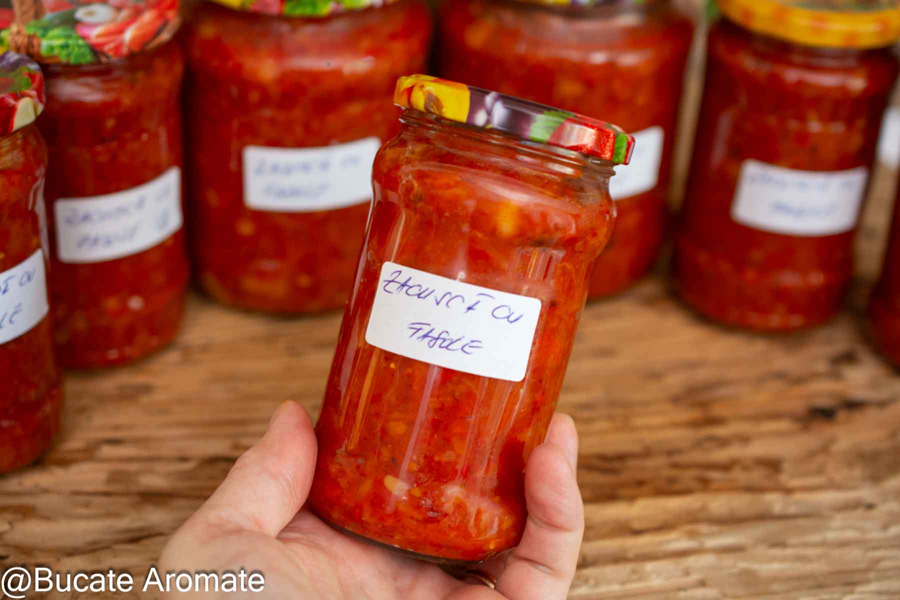 Borcan etichetat cu zacuscă de fasole boabe, ținut în mână, în fața celorlalte borcane