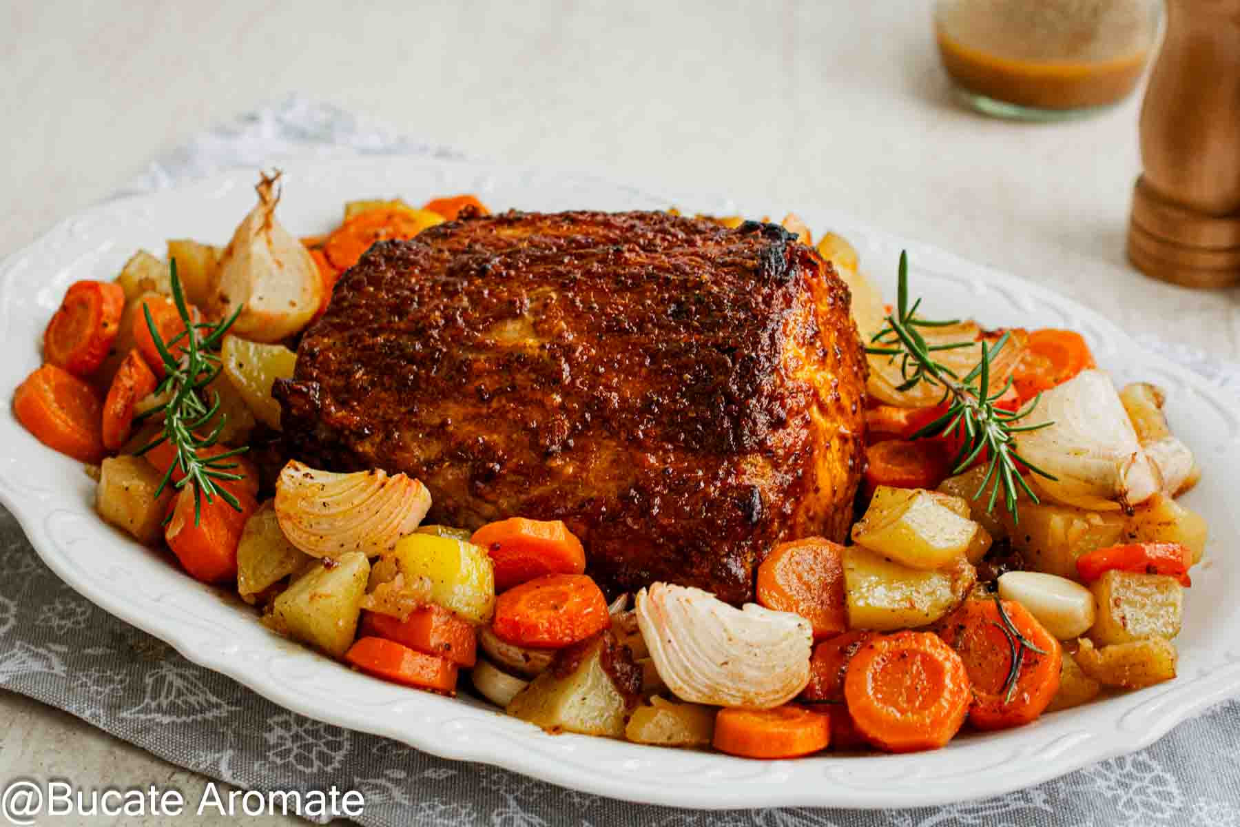 Cotlet de porc întreg, cu crustă brun-roșcată, pe un platou alb cu legume coapte și crenguțe de rozmarin.