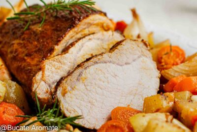 Prim-plan cu felii de cotlet de porc și marginea glazurată, alături de cartofi rumeniți, rondele de morcov și rozmarin.
