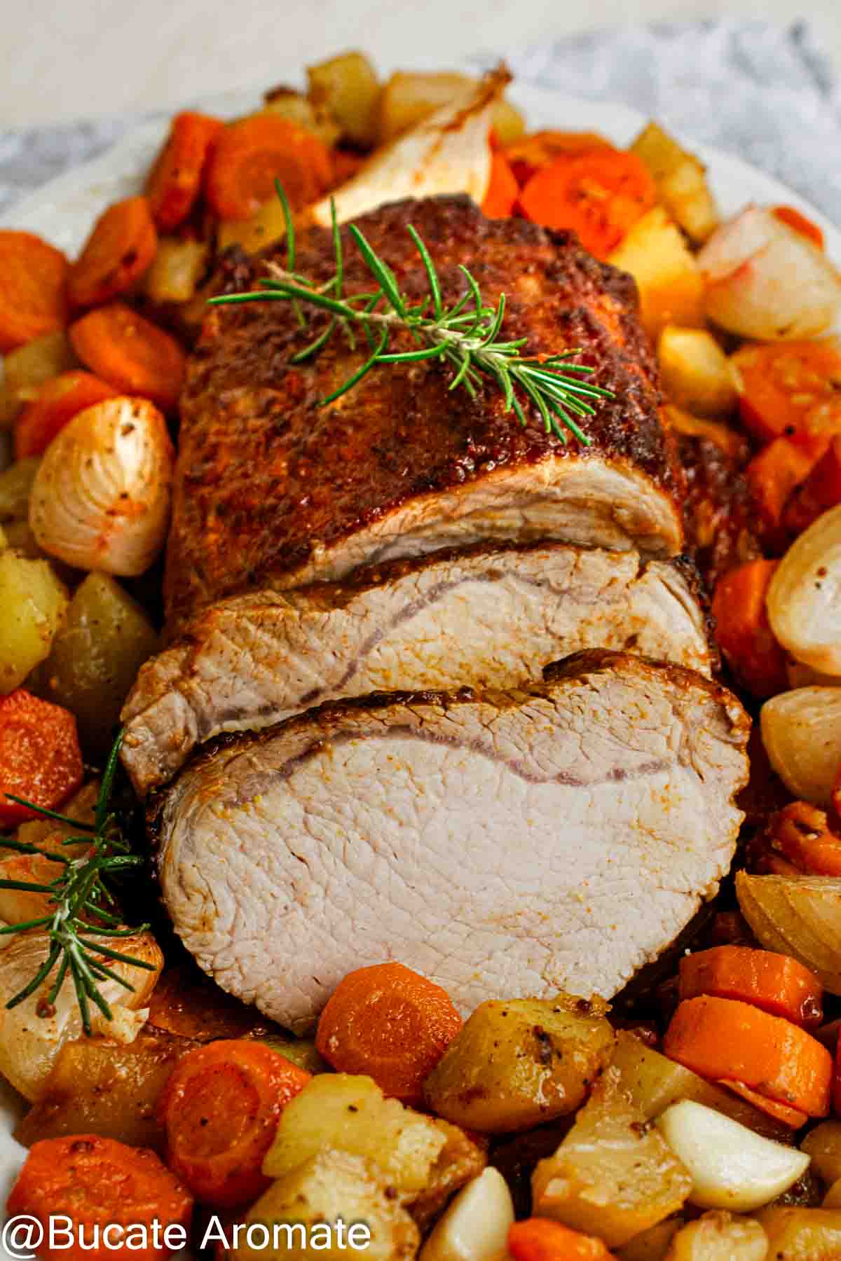 Cotlet de porc cu glazură rumenită, parțial feliat, înconjurat de cartofi, morcovi și ceapă la cuptor, decorat cu rozmarin.