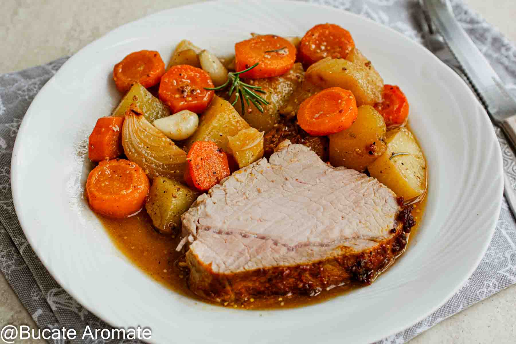 Felie de cotlet de porc glazurat, servită pe farfurie cu cartofi, morcovi, ceapă, usturoi și sosul fripturii.