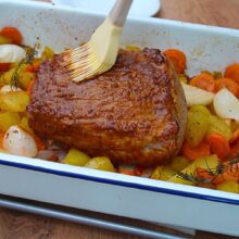 Aplicarea glazurii de muștar și miere cu o pensulă pe cotletul de porc, în tava cu cartofi, morcovi, ceapă și rozmarin.