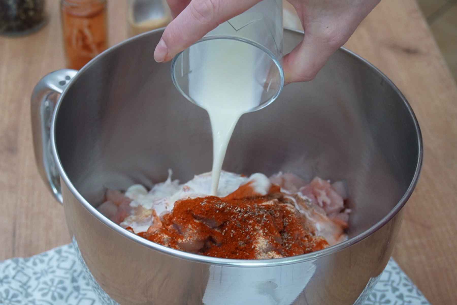 Smântână lichidă turnată peste amestecul de carne de pui, boia dulce, usturoi și piper, în bolul robotului de bucătărie.