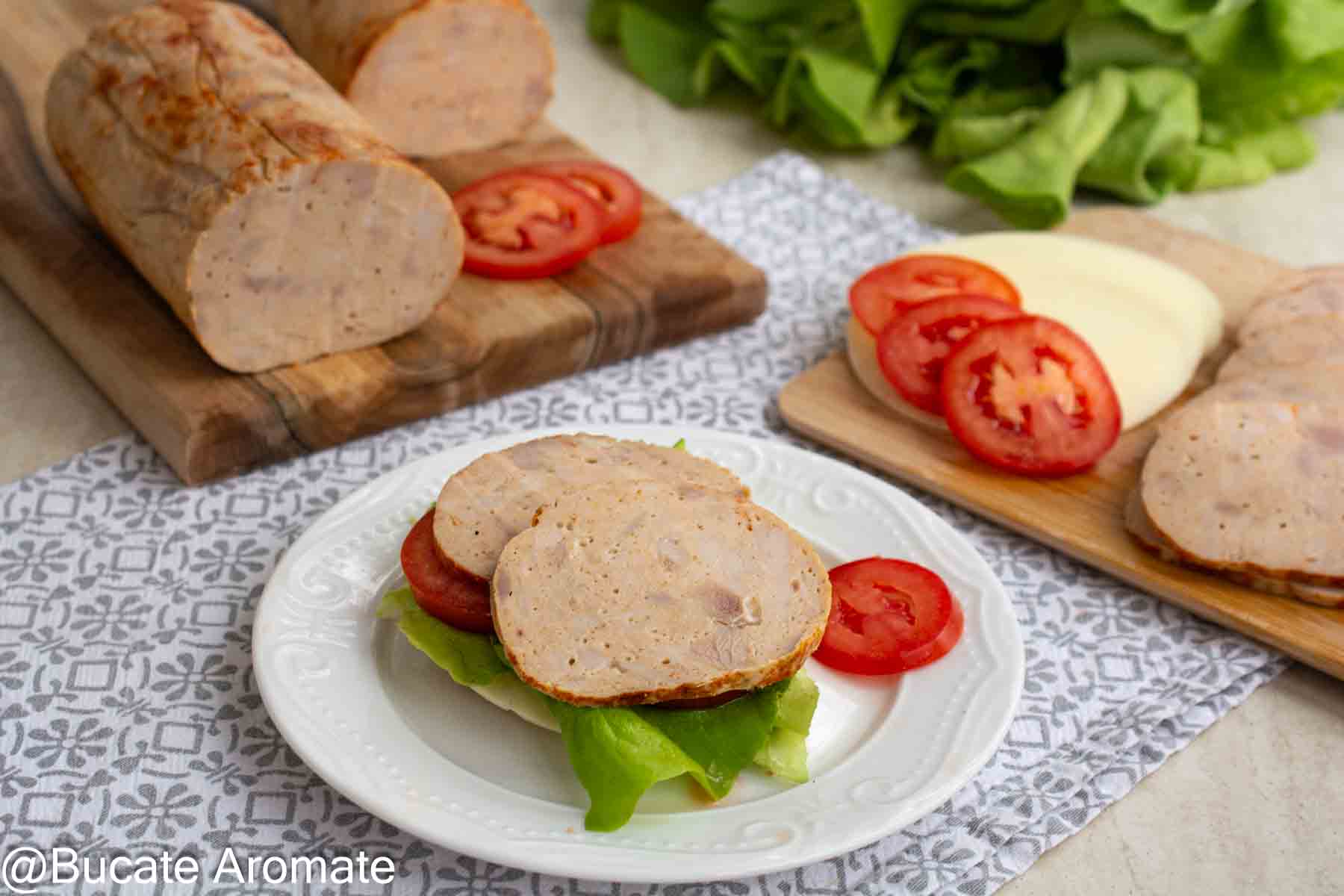 Sandviș cu șuncă de pui de casă, salată verde și roșii, servit pe o farfurie albă, cu batoane de șuncă feliată în fundal.
